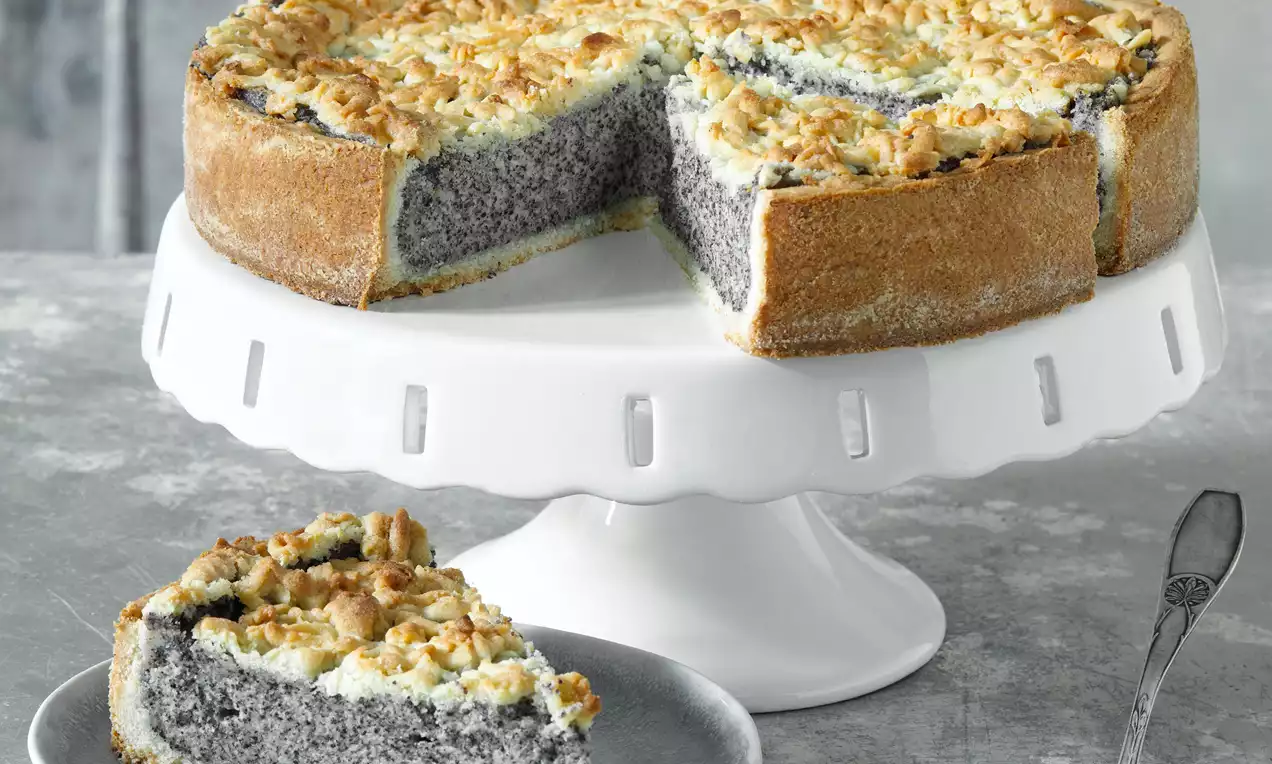 Makovo-tvarohový dort na bílé porcelánové míse s podstavcem, hrubší zlatavý okraj, světlý tvarohový povrch s drobenkou, šedý makový střed