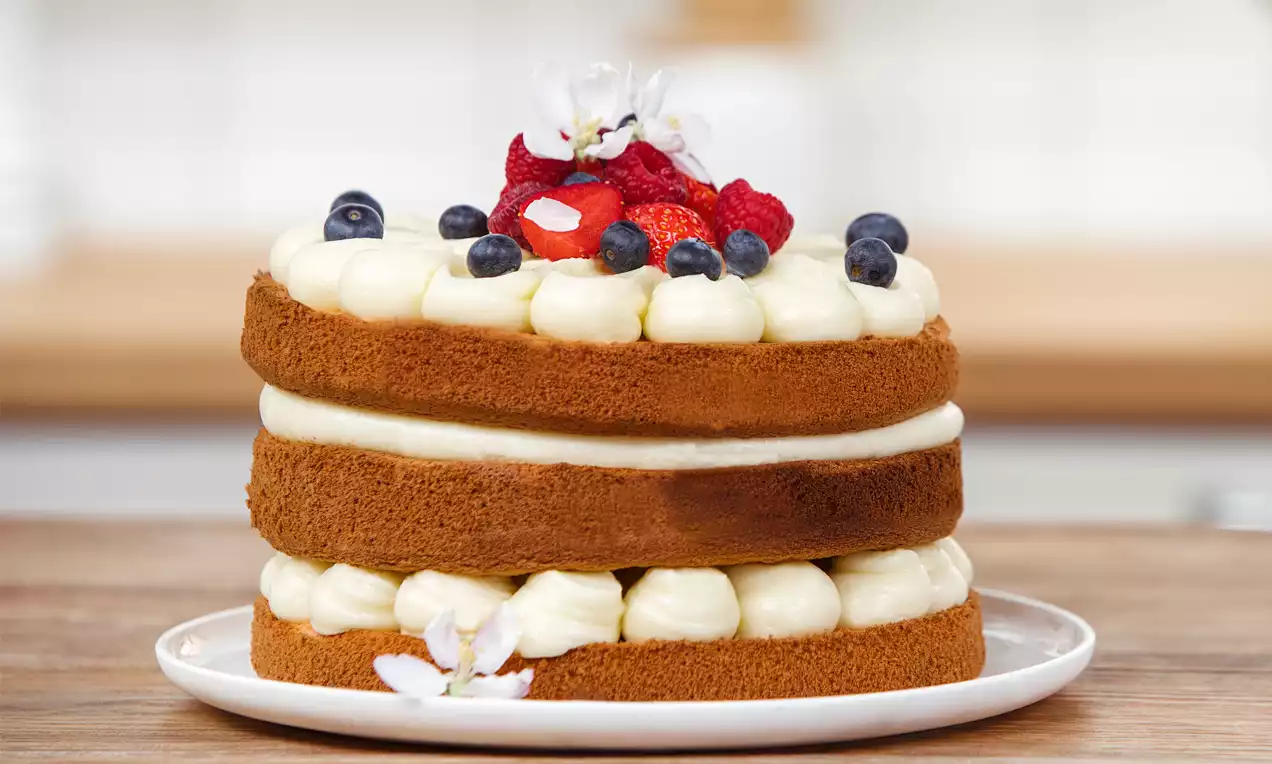 Naked Cake mit zwei goldbraunen Böden, heller Cremefüllung, frischen Beeren und Blüten dekoriert auf weißem Teller vor hellem Hintergrund