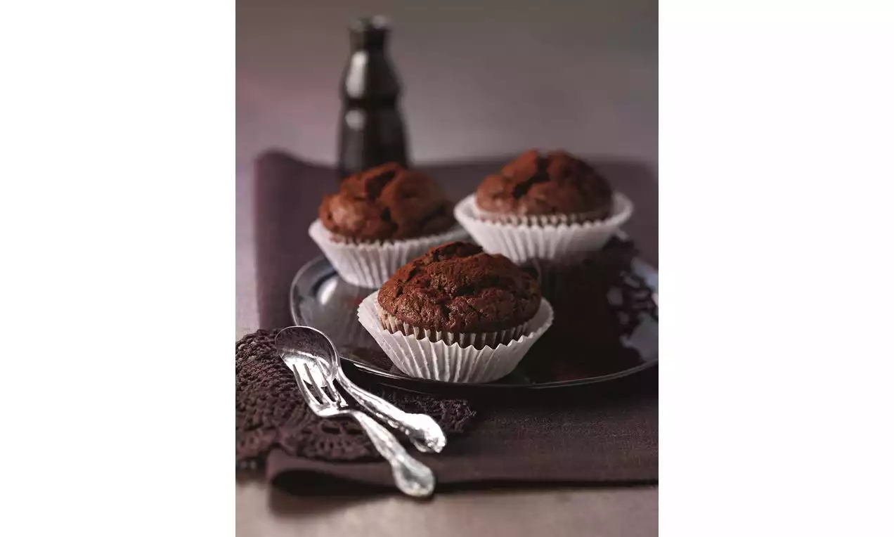 Donkerbruine chocolade-courgettemuffins in witte papieren vormpjes op een zwart bord, met zilveren lepels en een flesje op de achtergrond