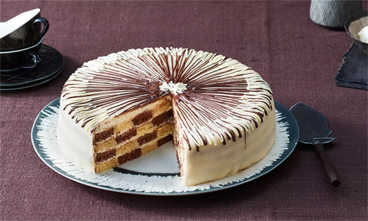 Кръгла шахматна торта със слоеве жълт и какаов блат, покрита с бяла глазура и декорация на тънки шоколадови линии върху голяма чиния