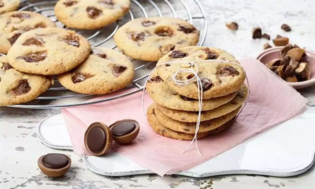 Zeltaini apaļi cepumi ar Toffifee gabaliņiem, sakrauti kaudzītē uz rozā papīra, līdzās izkārtoti konfekšu pusītes un izlobīti gabaliņi