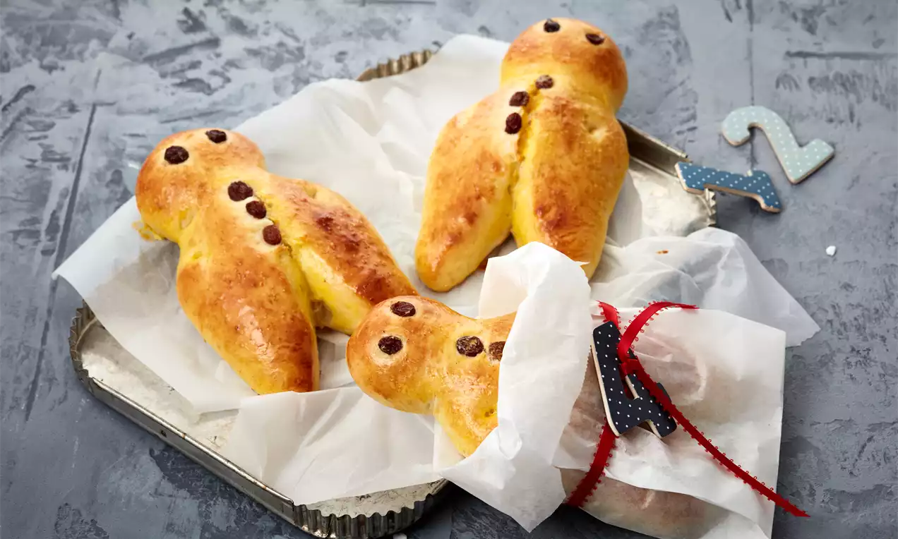 Panini dorati a forma di omino con occhi e bottoni di uvetta su un vassoio argentato foderato di carta, decorato con un nastro rosso