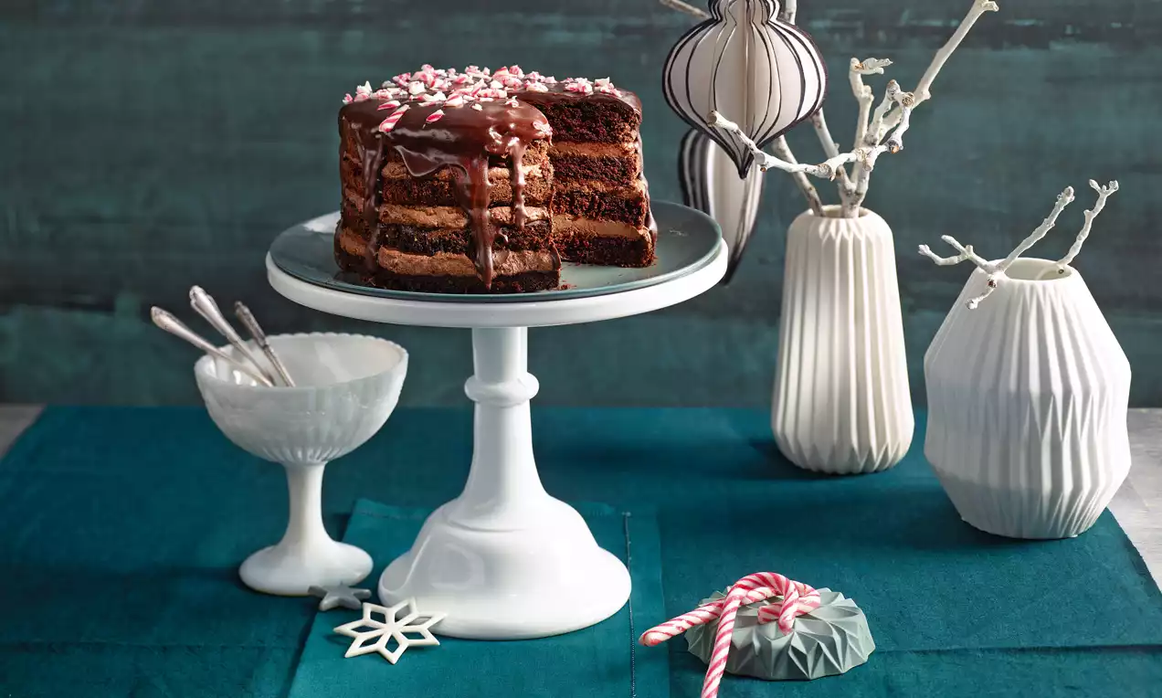 Dunkelbrauner Naked Cake aus saftigen Schokoböden und Cremefüllung, mit glänzender Glasur und gehackten Zuckerstangen auf weißer Tortenplatte