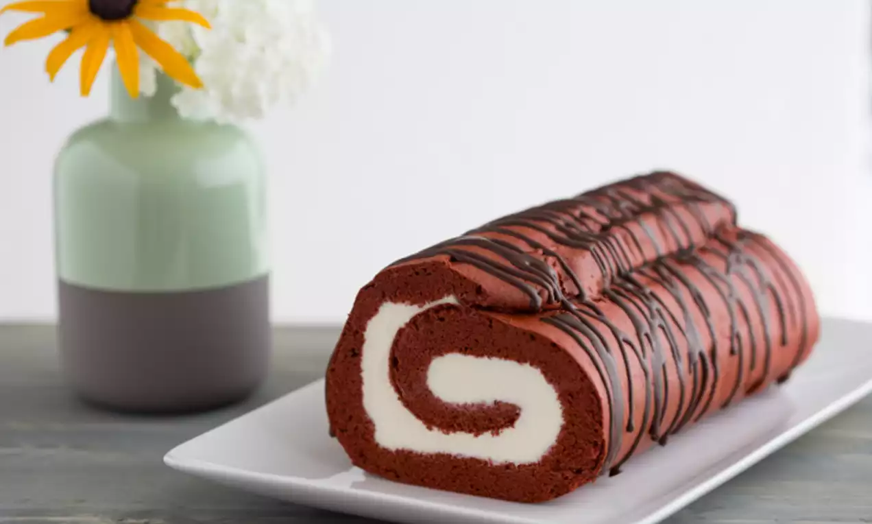 Rolcake van dieprode cake met een spiraalvormige witte crèmevulling, overgoten met donkerbruine chocoladeslierten op een wit bord
