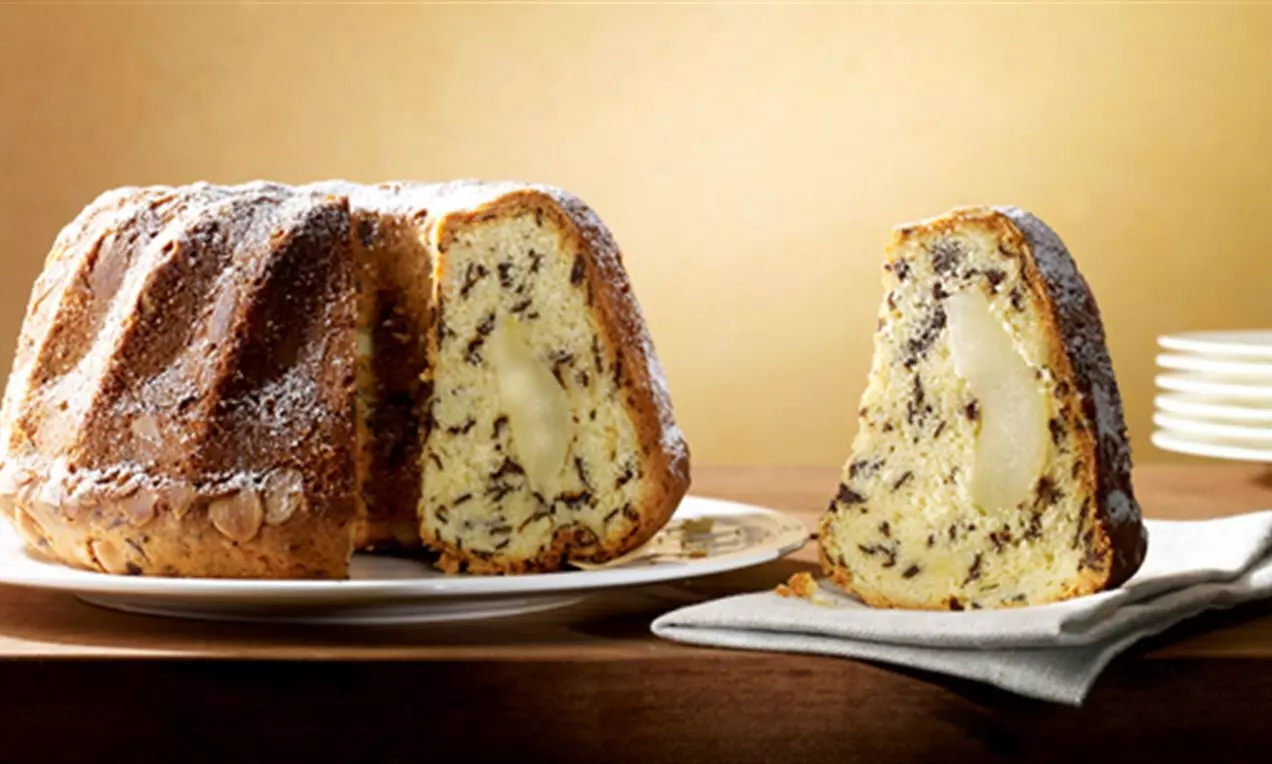 Златист кекс с шоколадови бучки и цели сочни круши, с леко поръсен пудра захар, сервиран върху бяла чиния на дървена маса