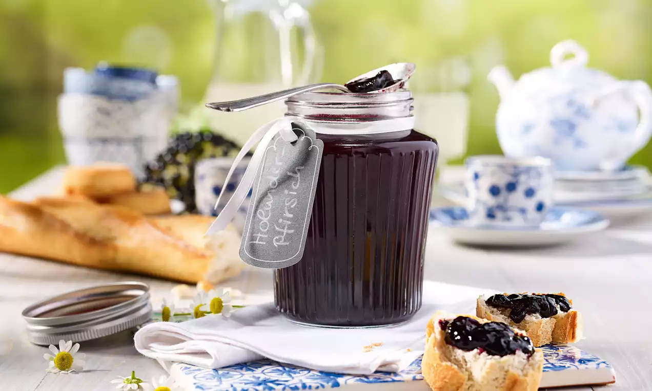 Geripptes Glas mit dunklem Holunder-Pfirsich-Gelee, beschriftetem Etikett und Löffel, daneben bestreichene Brotscheiben auf weißer Serviette