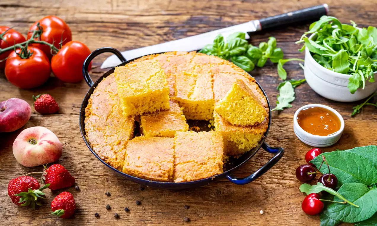 Goldgelbes Maisbrot in rechteckige Stücke geschnitten in einer dunklen Pfanne auf Holztisch, umgeben von Früchten, Salat und Messer