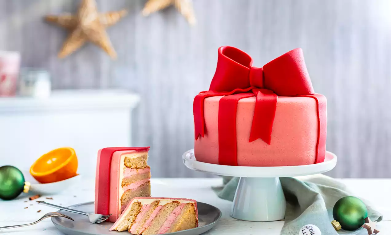 Okrugla torta u crvenom omotu od fondanta s mašnom na bijelom stalku, presjek s ružičastim slojevima i biskvitom na sivom tanjiru 