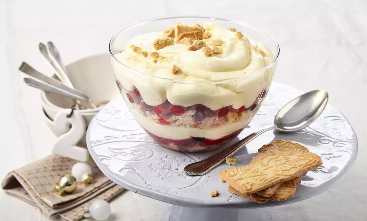 Schichtdessert in Glas mit heller Creme, roten Cranberries und Keksstücken auf dekorativer Tortenplatte aus weißem Porzellan