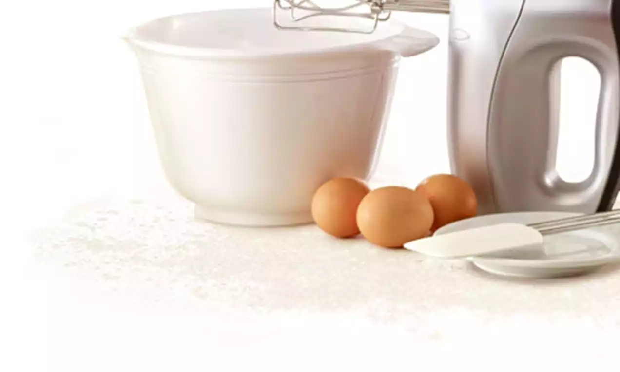 Witte mengkom met een metalen mixer op een lichte ondergrond, omringd door drie bruine eieren, een witte spatel en een helder bord