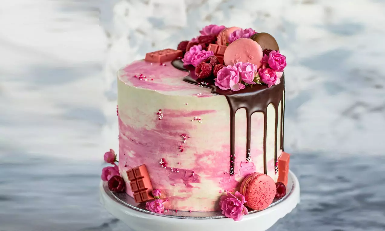 Висока торта с розово-бяла глазура, тъмнокафяви шоколадови струи, декорирана с малини, макарони, блокчета шоколад и розови цветя върху поставка