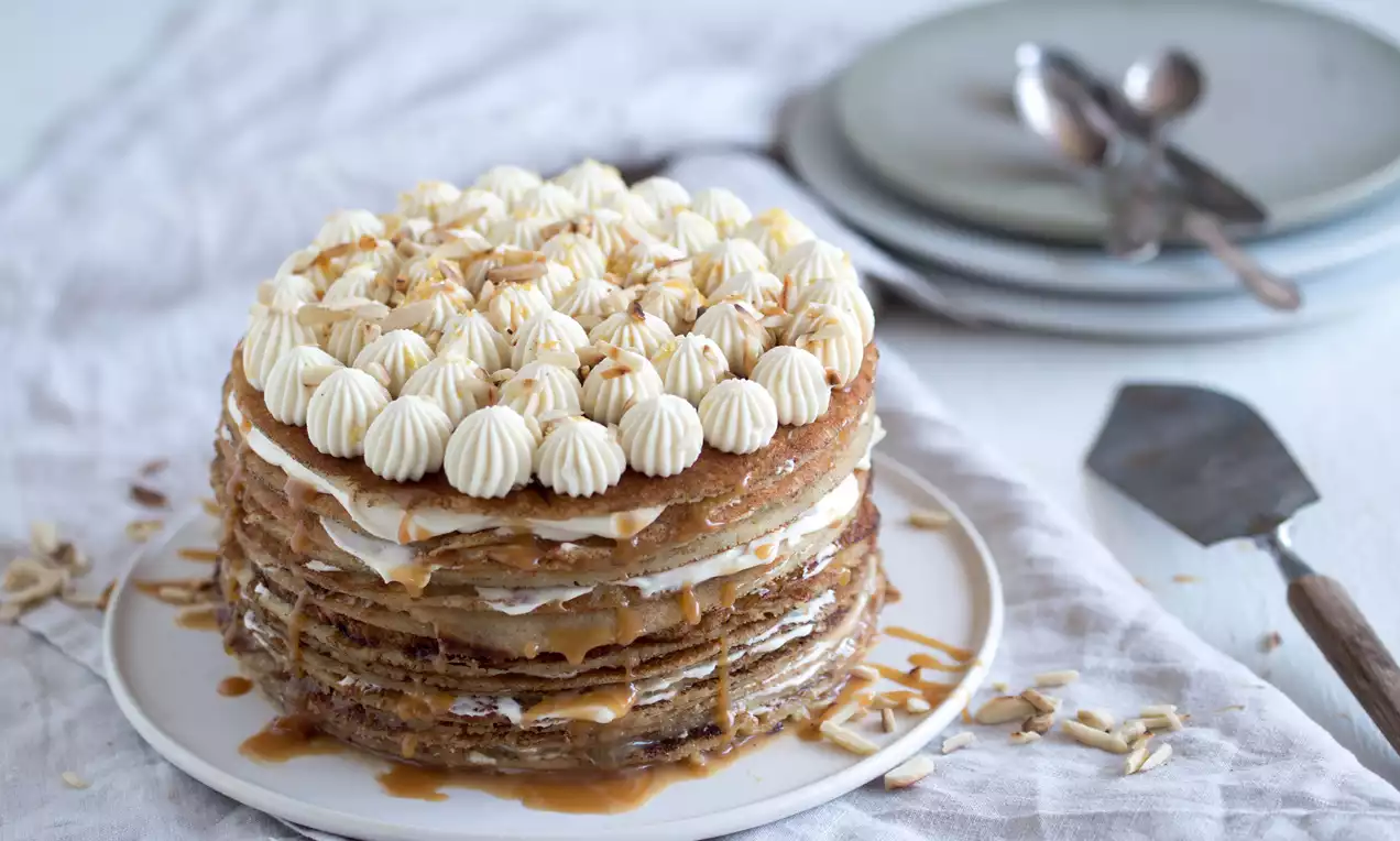 Pandekagelagkage med gyldne pandekagelag, lys flødeskum formet som små toppe og drys af nødder på en hvid tallerken