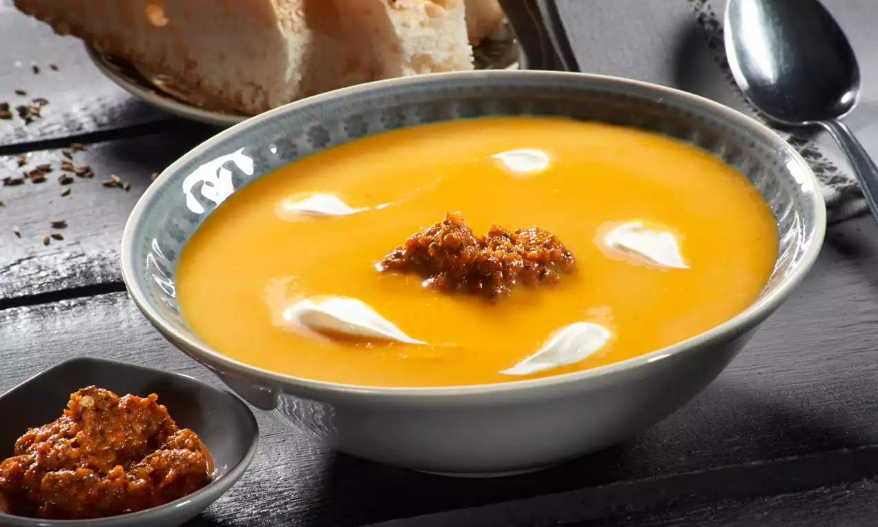 Svijetlonarančasta juha u keramičkoj zdjeli s hrskavom crvenkastom granolom u sredini, kremastim bijelim dijelovima i komadićima kruha u pozadini