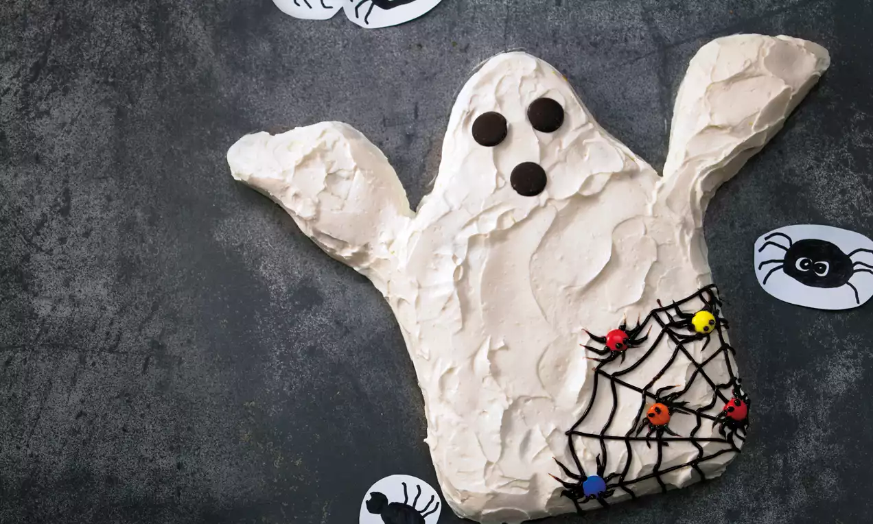 Ein flacher, hellcremer Gespenster-Kuchen mit Schokoladenaugen und Zuckerspinnennetz, dekoriert mit farbigen Zuckerspinnen auf dunklem Untergrund