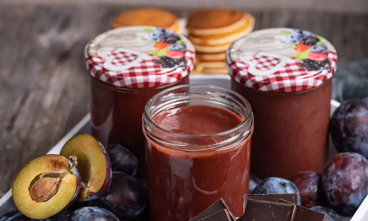 Svetleč kozarčki s sijočo bordo marmelado, obdani s svežimi modrimi slivami in koščki temne čokolade na lesenem pladnju