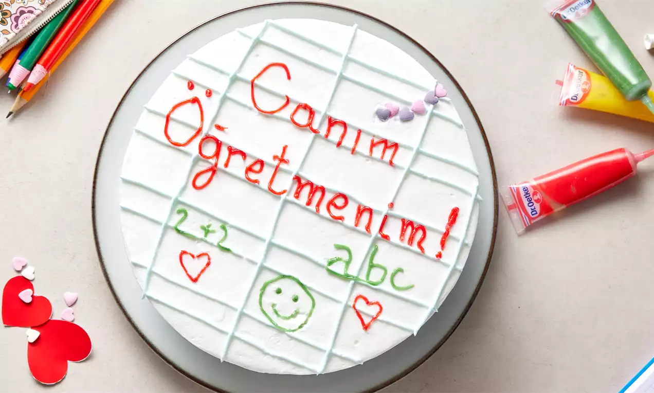 Beyaz kremalı yuvarlak pasta, üzerine renkli çikolata soslarıyla “Canım Öğretmenim!” yazısı ve çeşitli okul temalı çizimler yapılmış