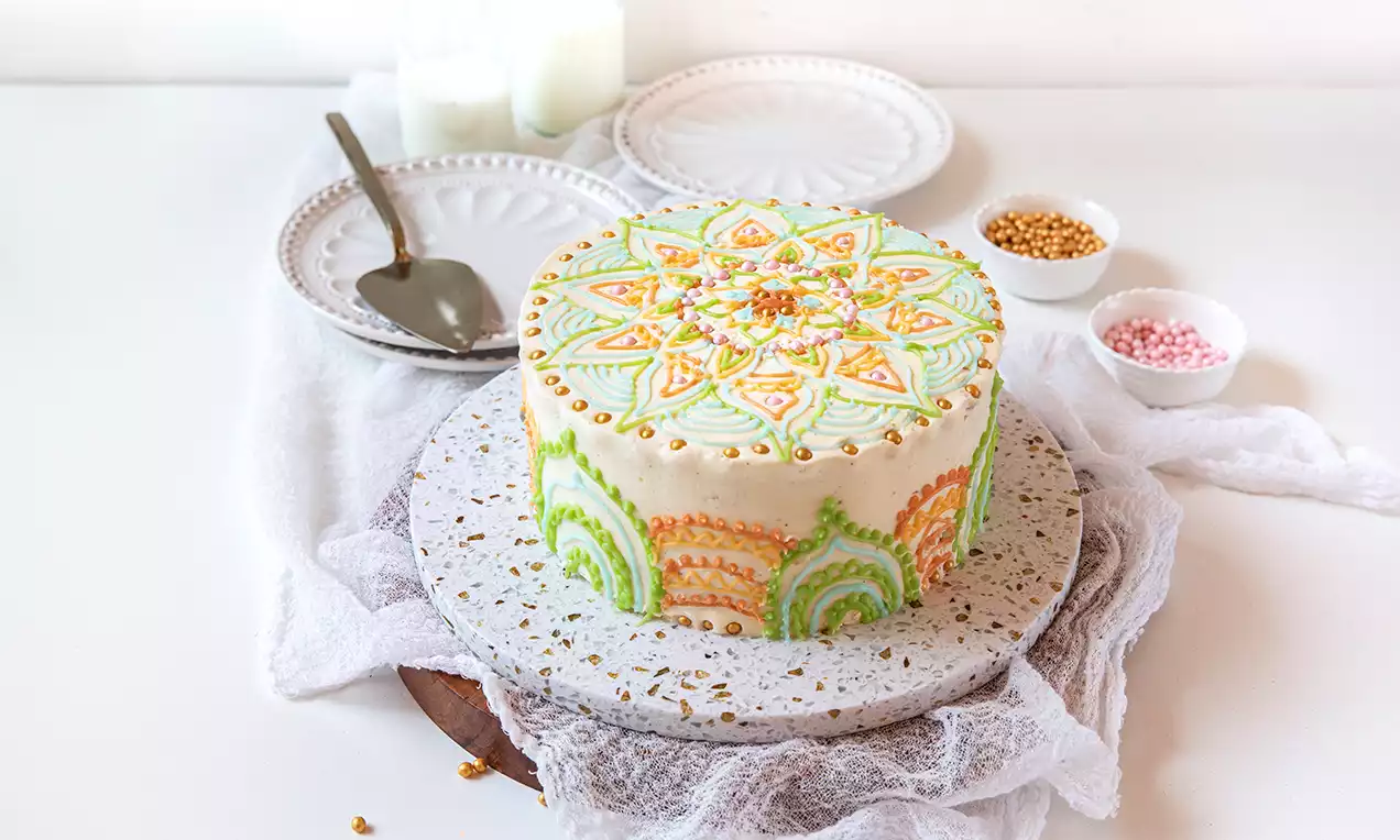 Korkea pyöreä kakku vaalealla kuorrutteella, käsin maalatuilla vihreillä ja oransseilla mandalakuvioilla, kultaisia helmiä koristeena marmorialustalla