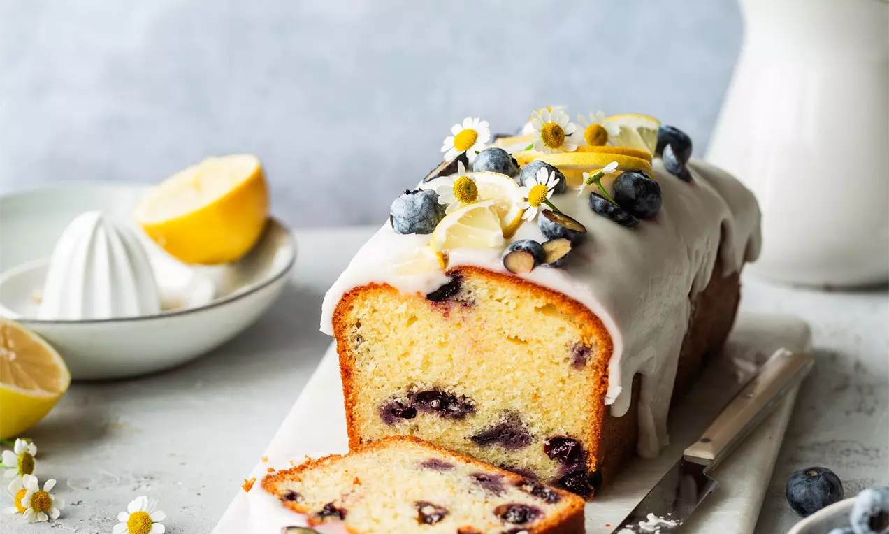 Picture - Golden blueberry loaf cake with white icing, topped with fresh blueberries, lemon slices, and small white flowers on a beige board