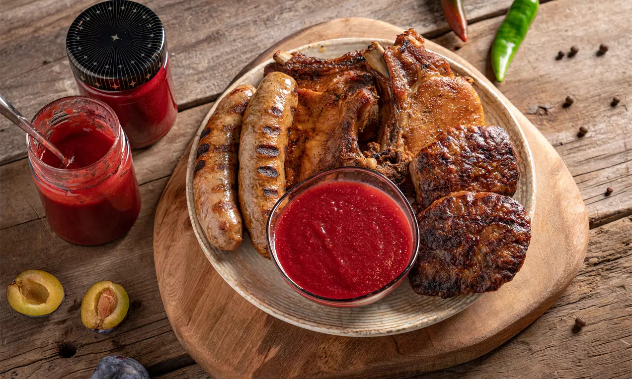 Гъст червен сос с гладка текстура в малка кръгла купичка, разположен върху дървена дъска до скарано месо и зеленчуци