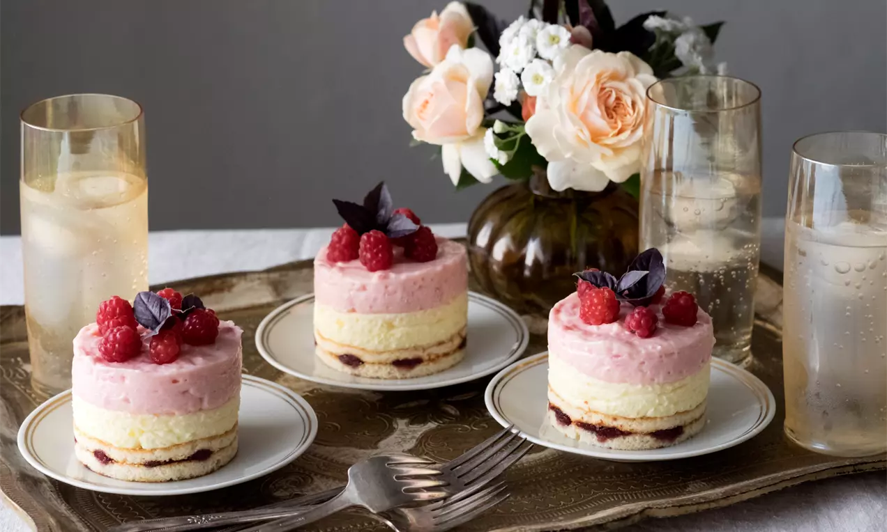 Кръгли слоести тортички с бледожълт крем, розов ягодово-малинов мус, гарнирани с пресни плодове и лилави листа върху порцеланови чинии