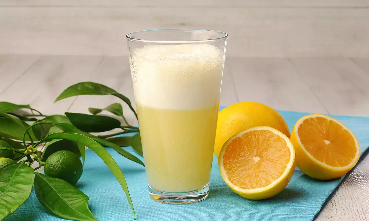 İnce uzun bardakta köpüklü limonata, yanında tam ve yarım limonlar, yeşil yapraklı dallar ve mavi örtü üzerinde düzenlenmiş