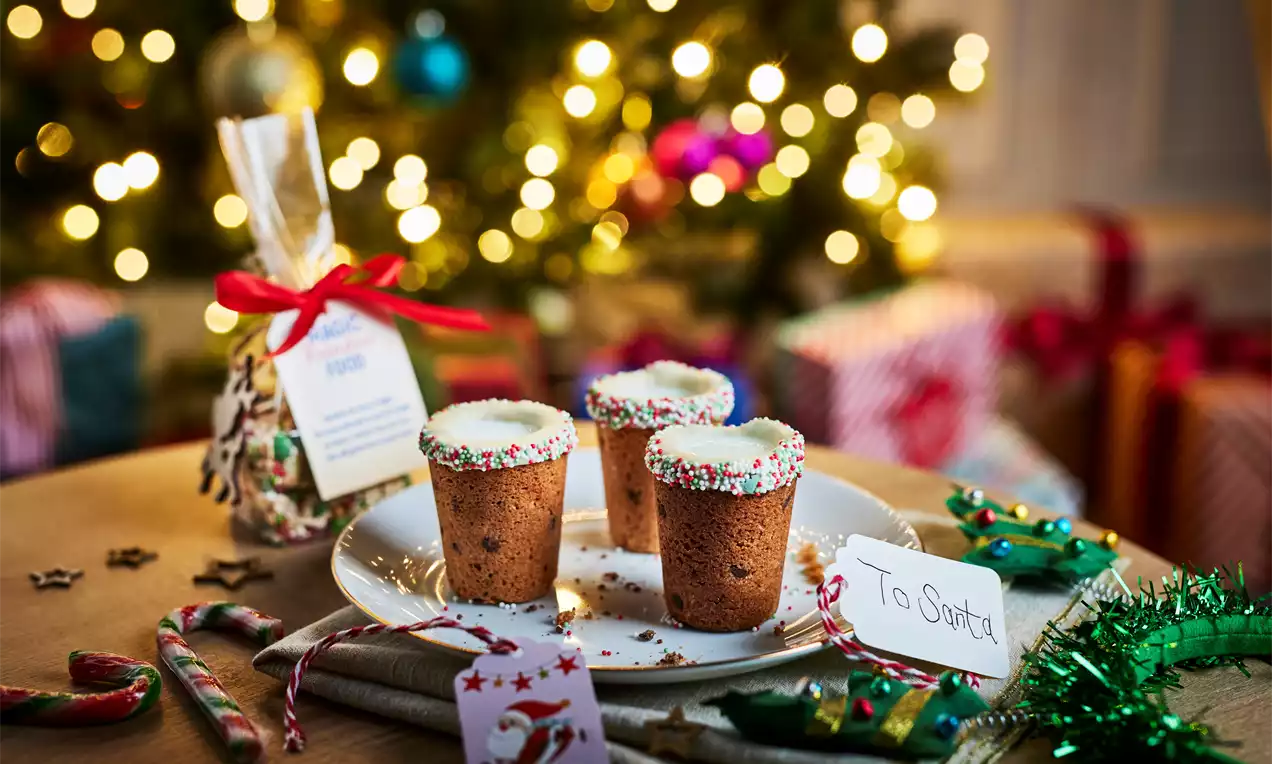 Brown cookie cups standing upright on a white plate, rims dipped in white icing and colourful sprinkles, with festive decorations nearby