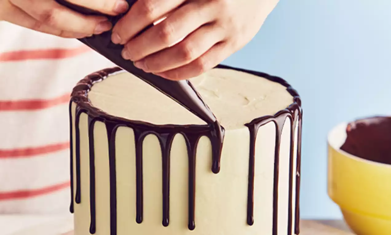 Chocolate ganache drips down the smooth pale cream frosting of a tall round cake, piped with precision against a blue background