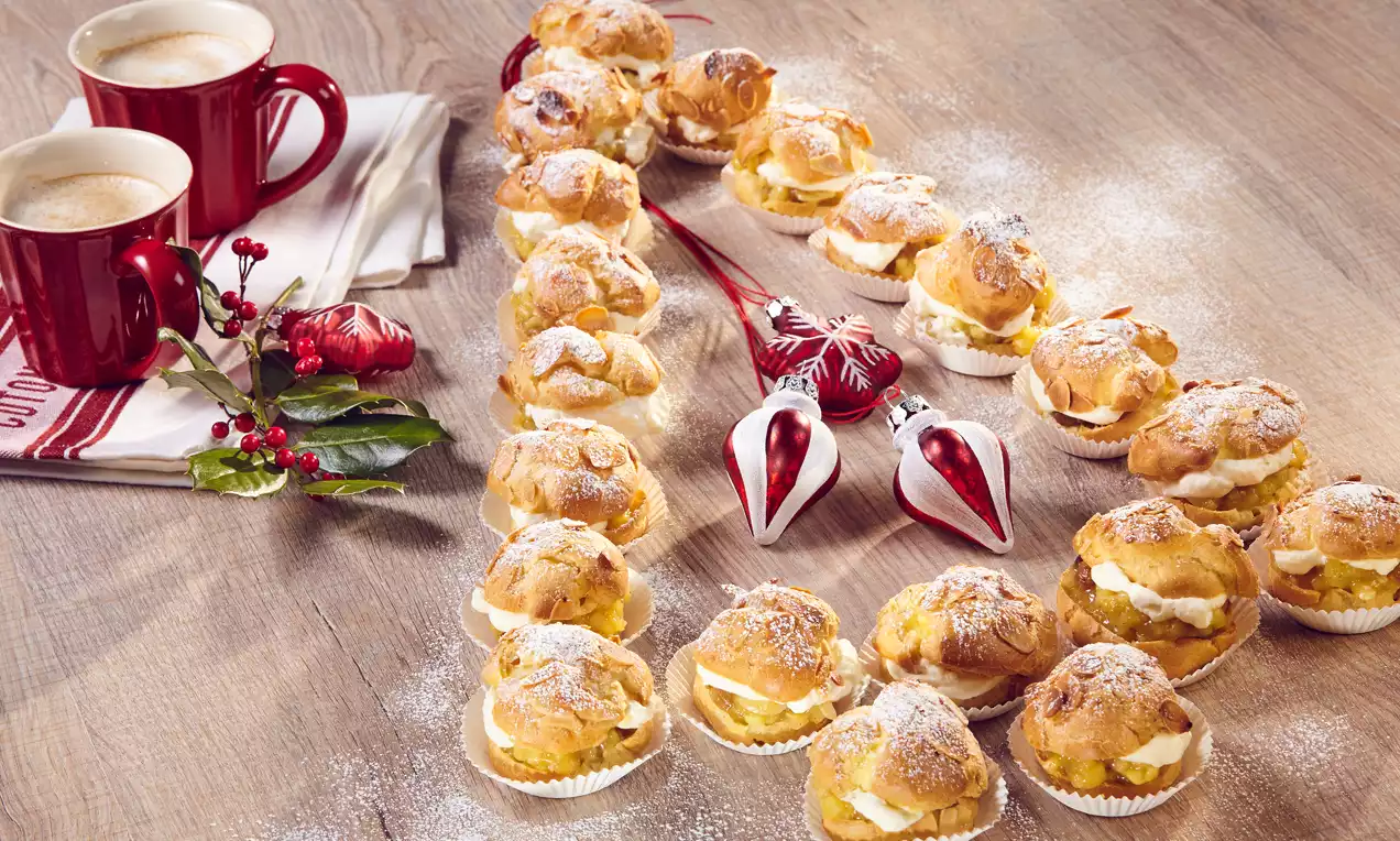Windbeutel in Tannenform mit goldbrauner Kruste, weißer Bratapfelfüllung, feinem Puderzucker, dekoriert mit roten Weihnachtsornamenten und Zweig