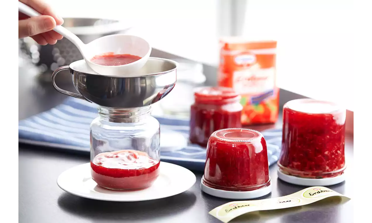 Rote Erdbeermarmelade wird mit einer weißen Kelle durch einen silbernen Trichter in ein Glas gefüllt, neben weiteren vorbereiteten Gläsern