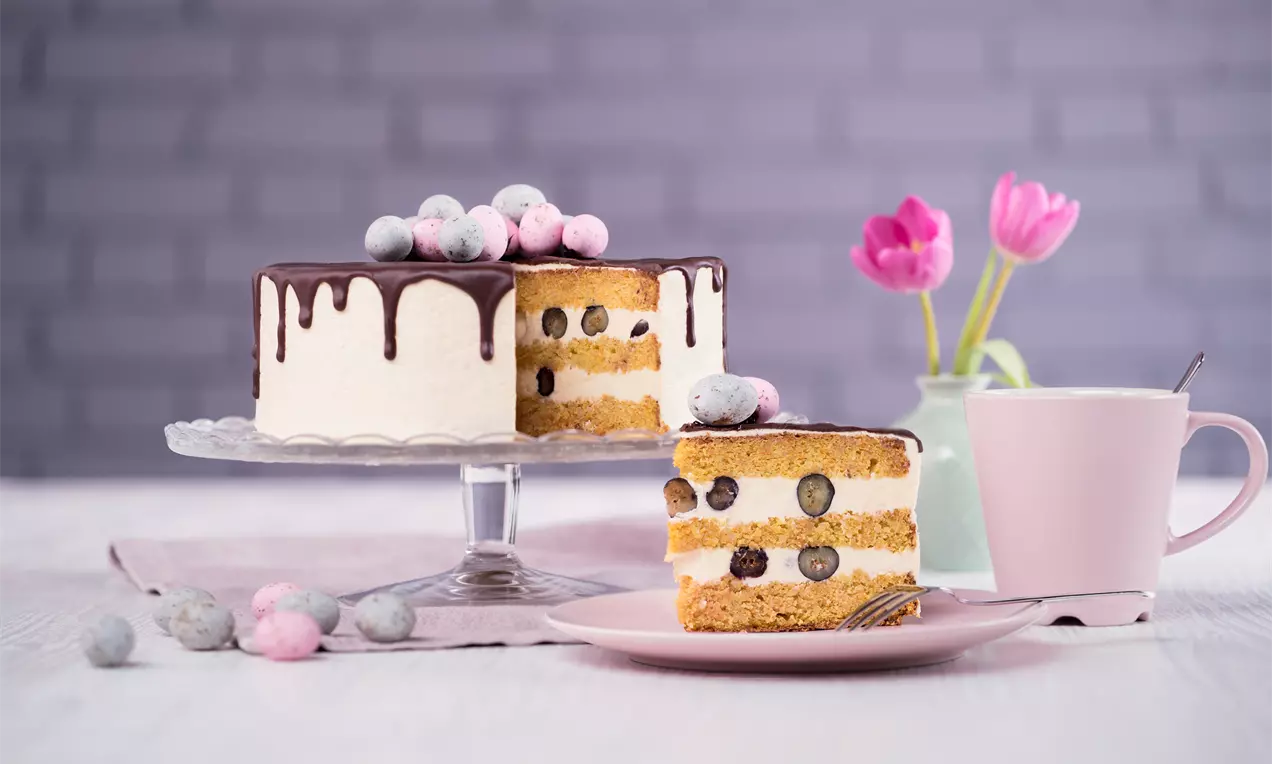 Кръгла торта с бели крем слоеве и боровинки, отгоре шоколадова глазура с цветни захаросани топчета, сервирана върху стъклена поставка