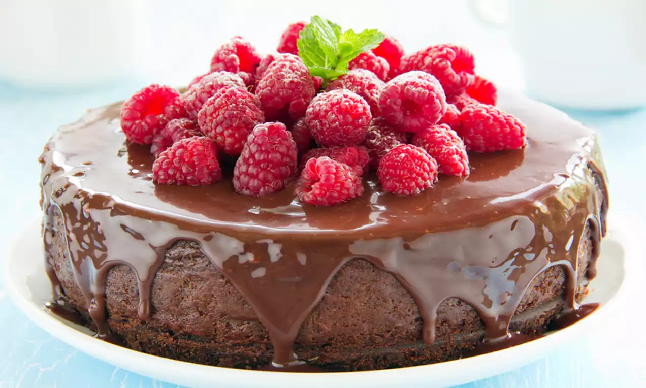 Chocoladecheesecake met glanzende donkere ganache, gegarneerd met sappige felrode frambozen en een groen muntblaadje op een wit bord