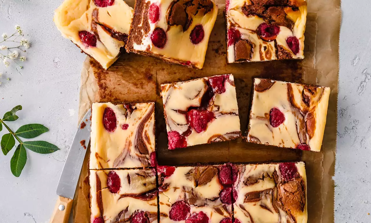 Picture - Square slices of vanilla cheesecake brownie with a golden base, marbled chocolate swirls, fresh raspberries, and rustic parchment