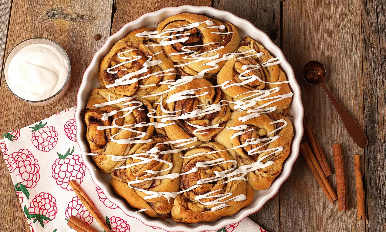 Tarçın şekerle kaplı altın rengi rulolar, beyaz krema sosu şeritleriyle süslenmiş, seramik bir tabakta ahşap zemin üzerine yerleştirilmiş