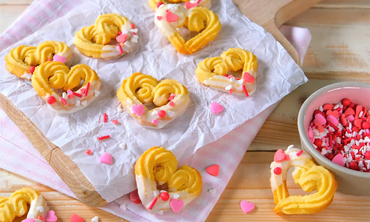 Spritzgebäck in Herzform, goldgelb mit weißer Glasur und rosa Zuckerstreuseln auf Pergamentpapier, daneben eine Schale mit Streudekor
