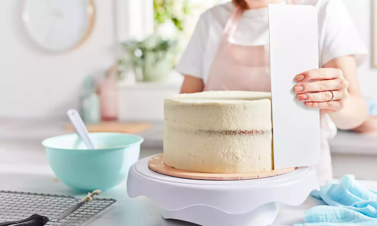 A round cake with pale buttercream being smoothed with a white tool on a rotating stand, set on a light countertop with baking tools nearby