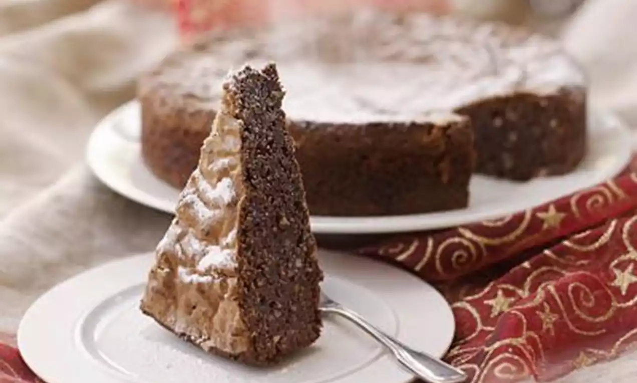 Mørk sjokoladekake med nøtter og dadler drysset med melis, servert som trekantet kakestykke på hvit tallerken med sølvskje, hel kake i bakgrunnen