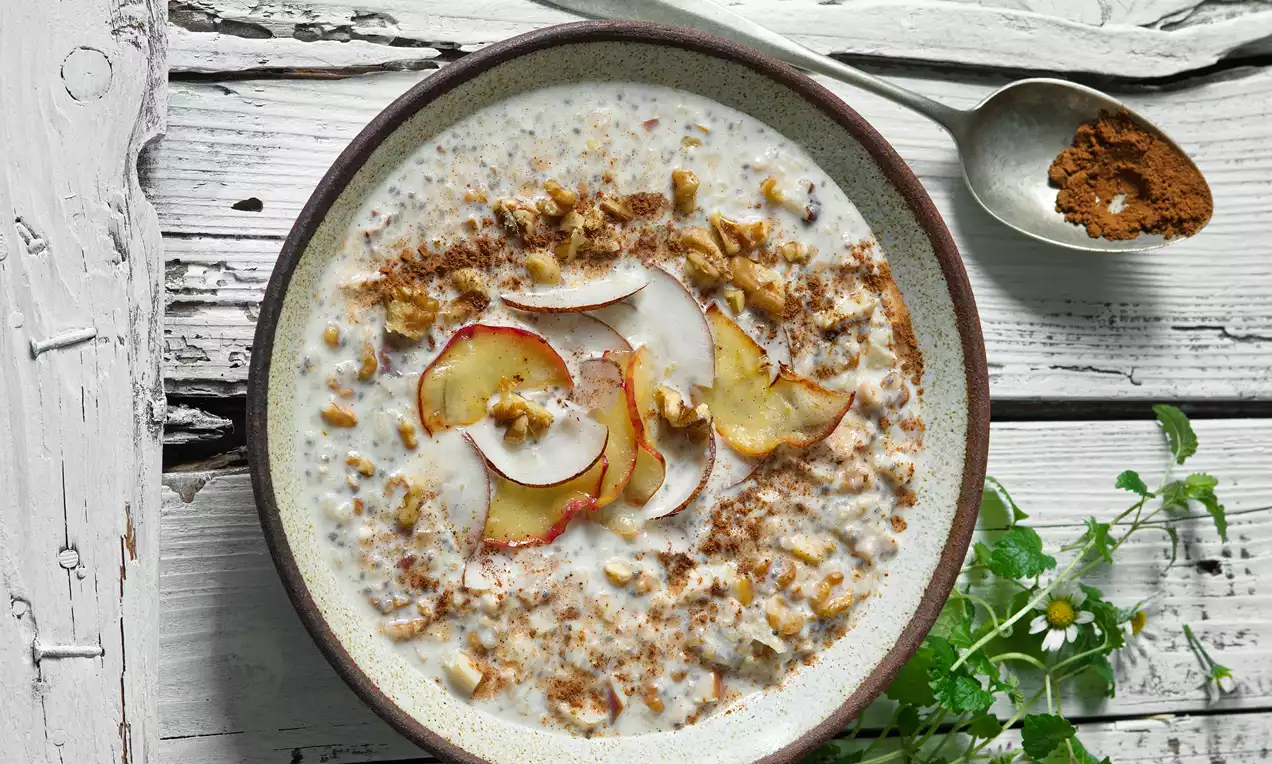 Fit rýžová kaše s jemně krémovou texturou, zdobená plátky jablek, sekanými ořechy a špetkou skořice na bílém dřevěném stole