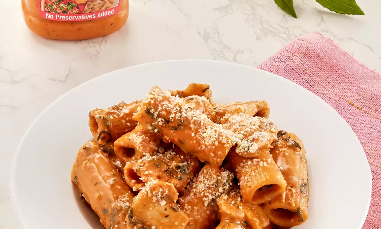 Rigatoni pasta in a creamy orange-hued sauce, sprinkled with grated cheese, served on a white plate atop a pink napkin