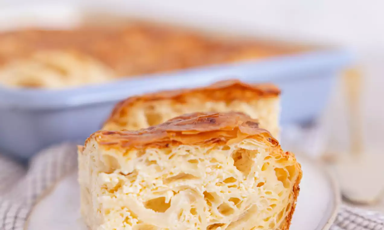 Dvobarvna rahlo zlata plastnata pita z nežnimi zračnimi plastmi na belem krožniku, ob podlagi iz svetlega blaga in modrem pekaču v ozadju