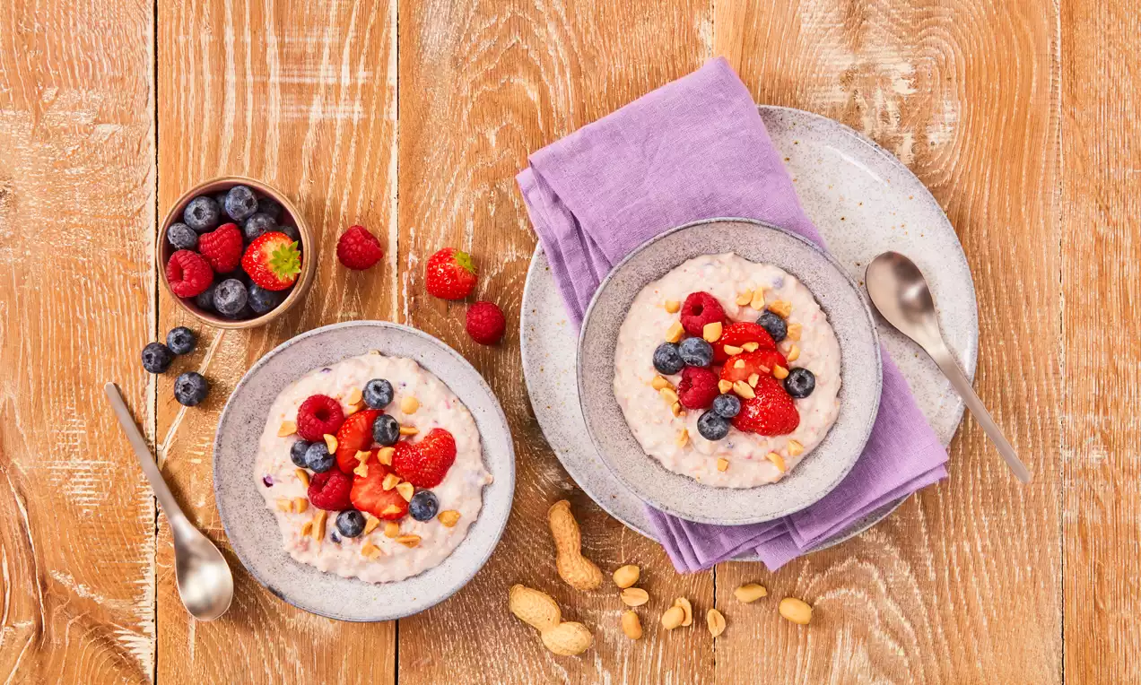 Cremiges, helles Proteinporridge in grauen Schalen, garniert mit roten Himbeeren, blauen Heidelbeeren und gehackten Erdnüssen auf Holzuntergrund