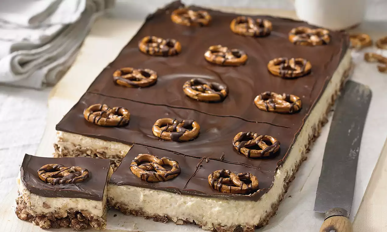 Carré croustillant à la base brun caramel, couche crémeuse beige, surface chocolatée décorée de bretzels brillants en cercle