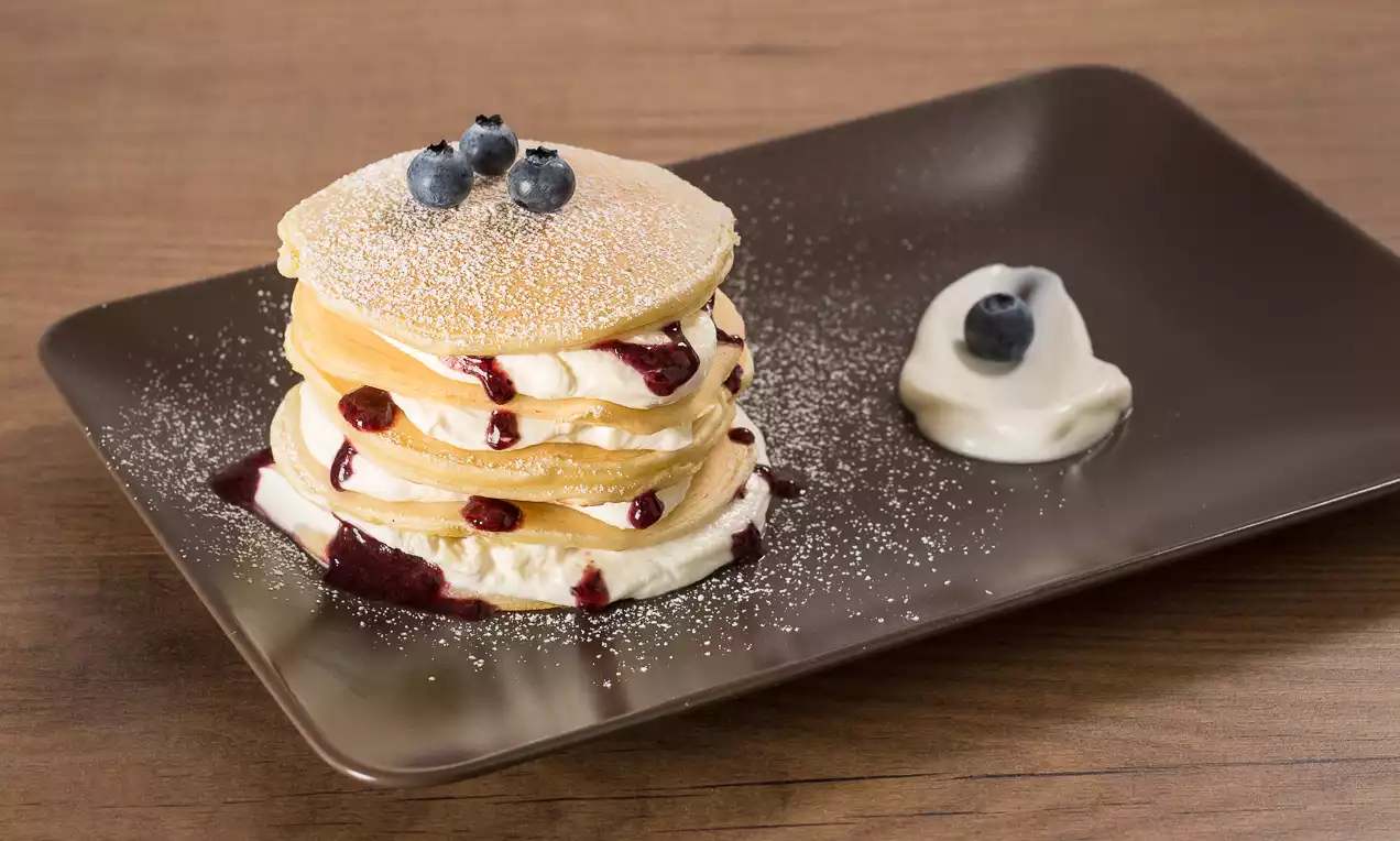 Pile de pancakes dorés nappés de crème blanche, coulis rouge et myrtilles, saupoudrée de sucre glace sur une assiette marron