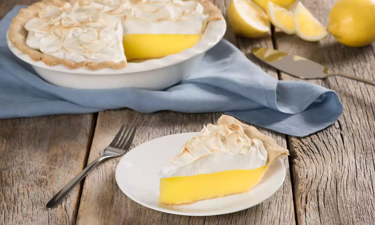 Lemon meringue pie with a vibrant yellow filling, toasted meringue peaks, and a flaky crust on a white plate beside a blue napkin and fresh lemons