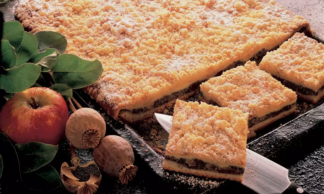 Goldgelber Mohnkuchen mit knusprigen Streuseln, in Stücke geschnitten auf einem dunklen Blech, dekoriert mit Mohnkapseln und einem roten Apfel