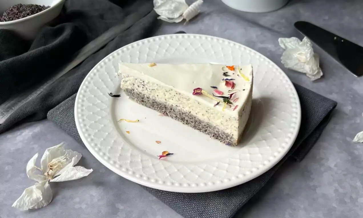 Makový cheesecake s vrstvenou texturou, bílou krémovou náplní, posypaný květy na zdobeném talíři položeném na tmavé látce