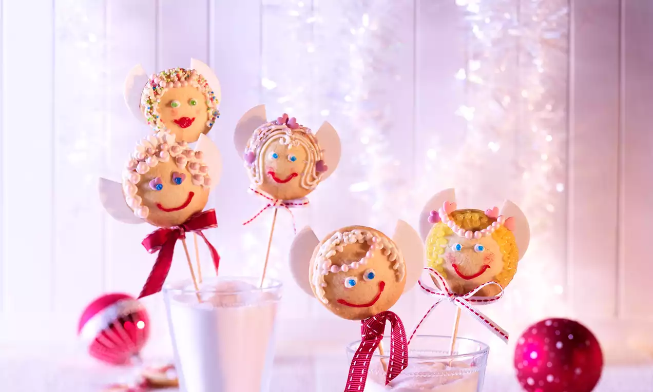 Sucettes rondes dorées décorées en anges avec ailes blanches, visages souriants et rubans rouges, placées dans des verres sur fond festif