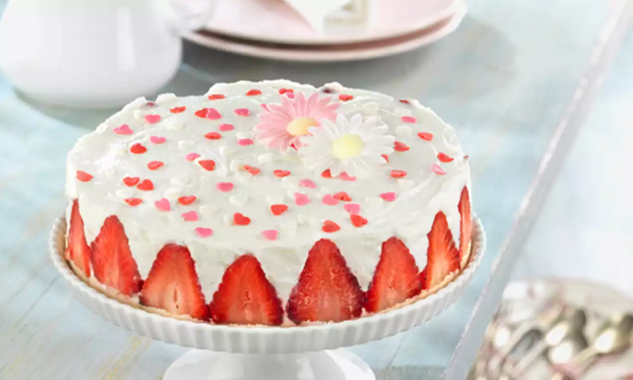Tarta de yogur blanca decorada con fresas cortadas en los bordes, confeti de corazones rojos y rosas y flores de azúcar sobre un plato elevado