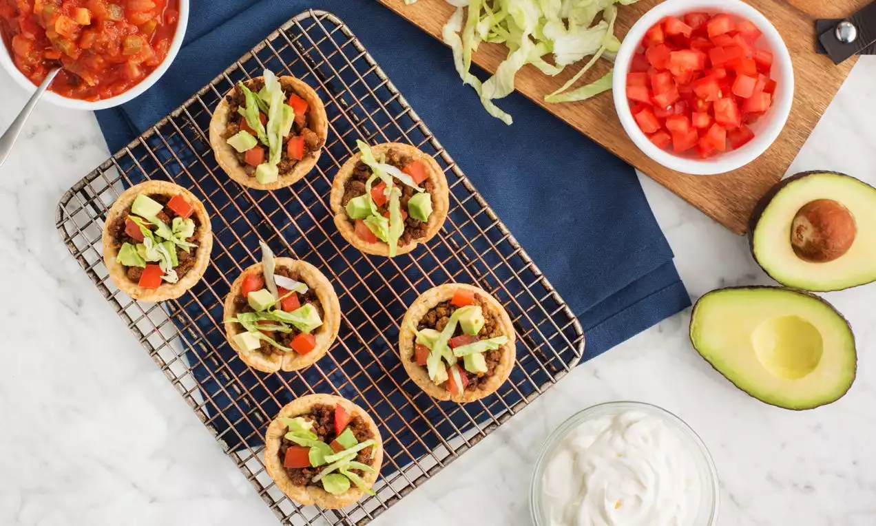 Mini taco cups with golden crispy shells, topped with diced red tomatoes, green avocado pieces, shredded lettuce, and melted cheese on a wire cooling rack over a dark blue cloth, surrounded by bowls of salsa, sour cream, and fresh avocado halves