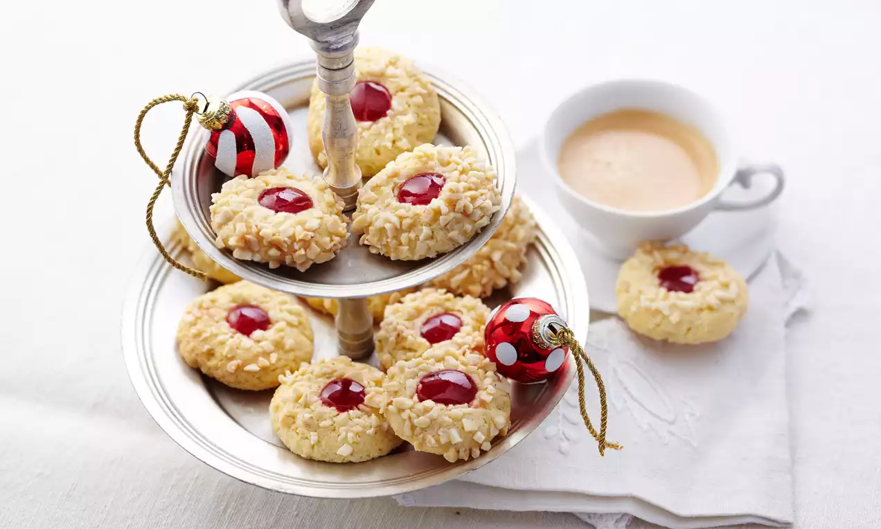 Helle Kulleraugen mit roten Marmeladenfüllungen und gehackten Nüssen auf silberner Etagere, daneben eine Tasse Kaffee auf weißem Stoff