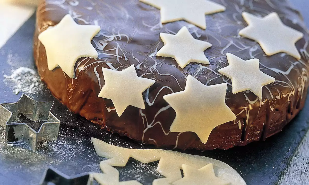Schokoladentorte mit glänzender Glasur und dekorativen weißen Sternen aus Marzipan auf einer dunklen Kuchenplatte mit Sternförmchen